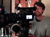 A person looks through the viewfinder of a big digital film camera while two people stand behind them looking at what the camera is filming.