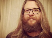 Faculty member Chris Faulker with a full beard, long hair, and glasses looks into the camera in sepia lighting.