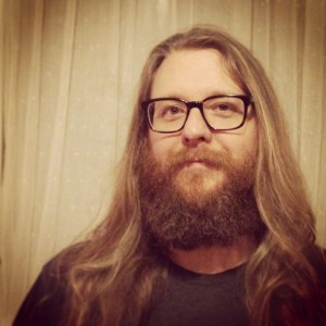 Faculty member Chris Faulker with a full beard, long hair, and glasses looks into the camera in sepia lighting.
