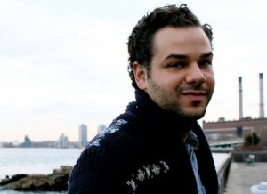 Profile picture of NElson Isava as they stand outside near a body of water looking into the camera, wearing a jacket and button up shirt.