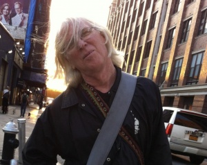 Profile photo of faculty member Bill Hopkins taken outside a busy street. A bag strap hangs across his black jacket as he looks into the camera with a sun setting behind him.