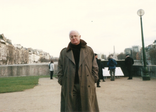 David Black, wearing a trench coat, stands outside in front of a body of water looking directly into the camera with people behind him in the background.
