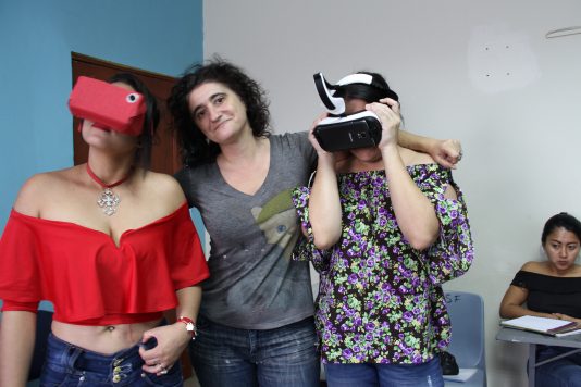 Alessandra Zeka stands in the middle of two people, her arms affectionately around both of them. The two people on either side of Alessandra are both in the midst of using the occulous VR headset, looking in different directions as they experience the virtual reality.