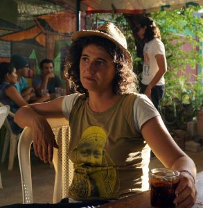 Alessandra Zeka sits in a chair under a text holding a drink in her left hand, resting it on a table. A group of people converse behind her.