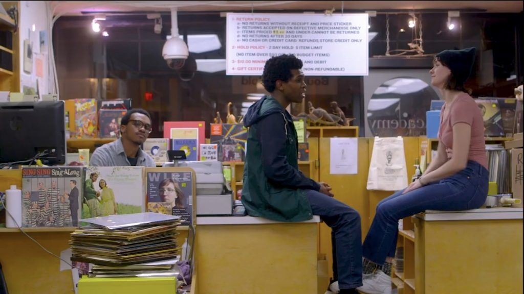 Lanre sits in a book store looking bored while two patrons sit on the counter conversing, seeming to pay him no mind.