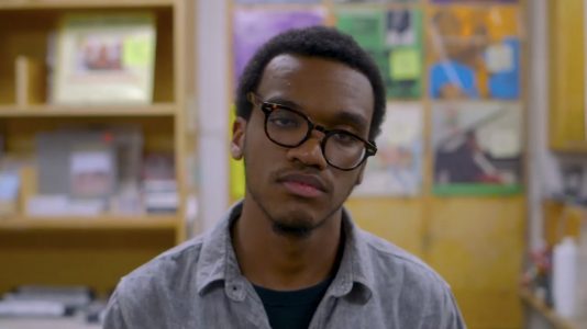 Profile photo of director Lane from a still from his film. He looks direclty at the camera while in a bookstore wearing black glasses.