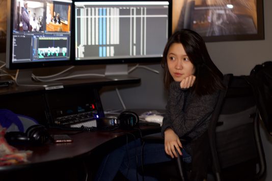 Charlotte Atkinson sits at the SVA editing lab Computer workstation looking at an editing timeline on the computer with her head resting on her hand.