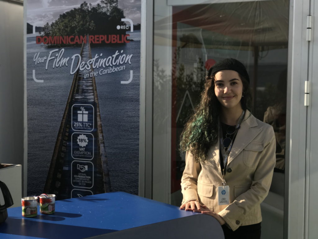 Filmmaker Paula Curry stands in a beige blazer outside in front of a poster that says "Dominican Republic Your Film Destination in the caribbean" While smiling at the camera.