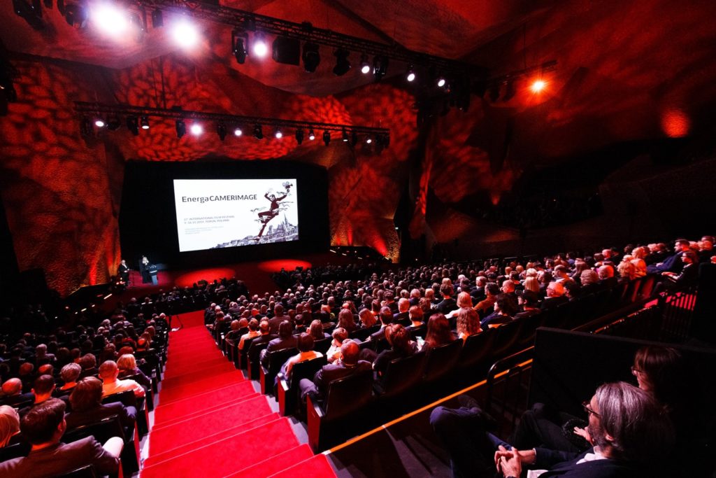 A packed auditorium of people sitting watching the screen that has the word "energaCameraimage" on it. The auditorium is all red.