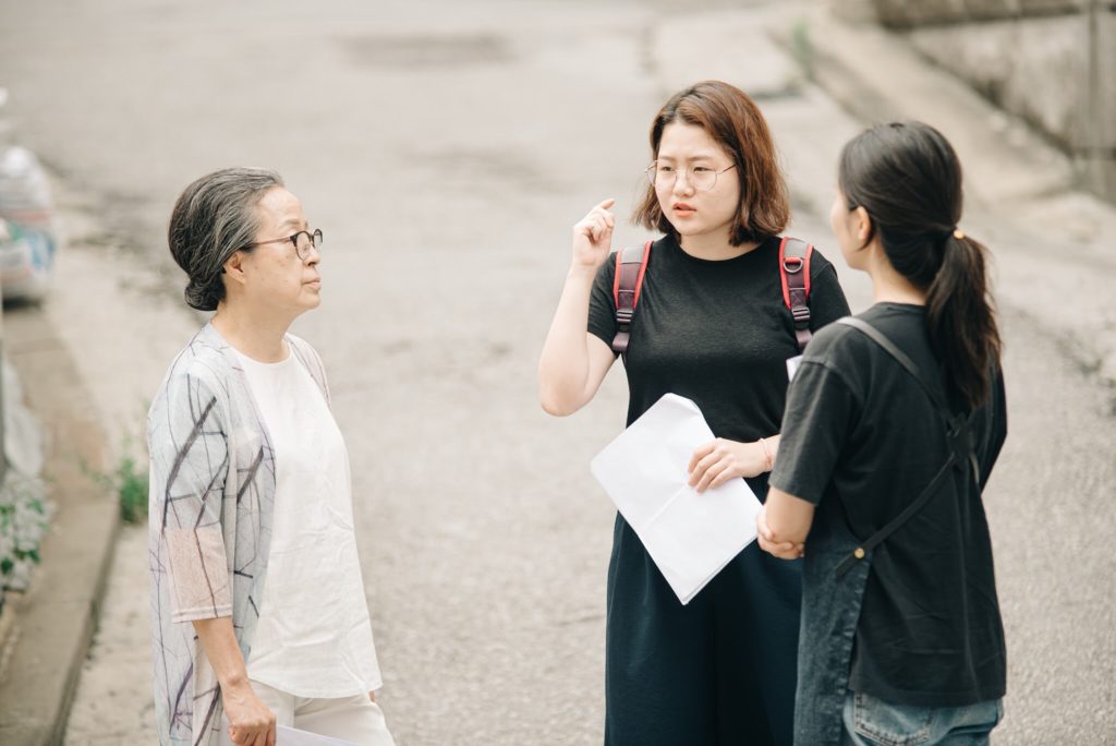 Three people stand outside talking to eachother. one of them has a backpack on and is holding a screenplay. Another wears a black shirt with their back to the camera and the third person looks at them while wearing a white shirt and a gray cardigan.