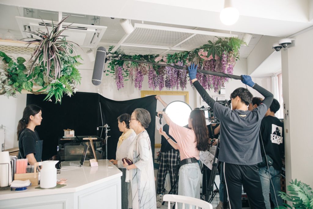 Behind the scenes of a a shoot in process. A person stands at a cafe counter looking at the cashier while a film crew stands to the side filming them. 