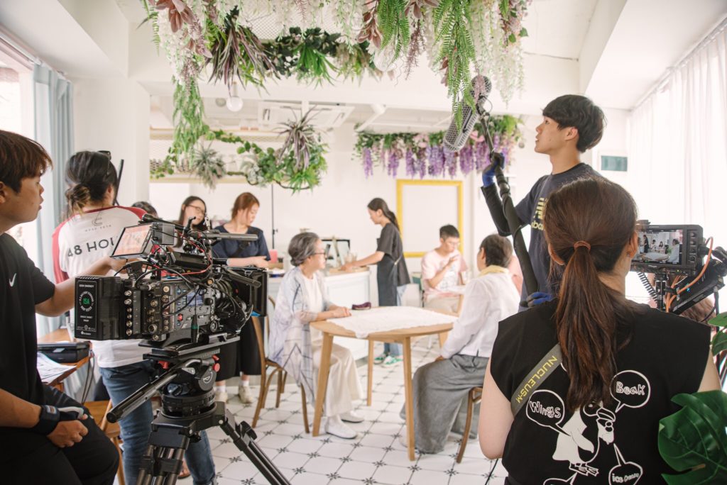 Behind the scenes shot of SVA students  Jungyoon Jang & Hae Seoung Kim film. A person with a camera films restaurant patrons eating inside an all white cafe that has hanging plants decorating the ceiling. a person with a boom pole stands to the side while another person holds a viewfinder on the right.. 