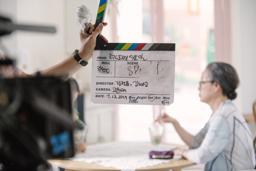 A hand holds a camera slate ready to clap before filming beings. Behind it, an actor sits at a table slightly out of focus.