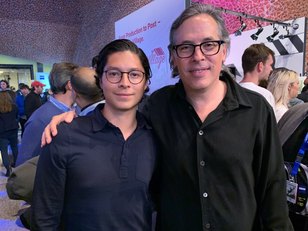 Alejandro cortes wears a black long sleve polo next to a person with a black button up shirt. both are smiling while a crowd of people stands behind them, but not paying them any attention.