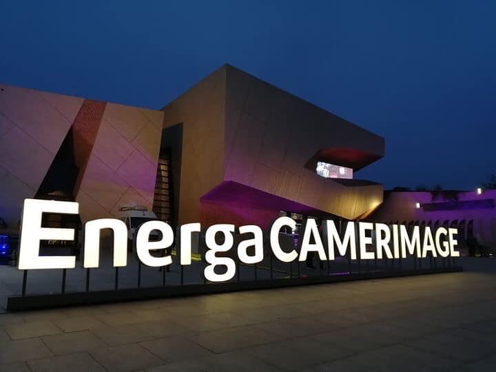 A building with the word "EnergaCAMERAIMAGE" in big letters as a sign outside of it. Purple lights shine on the building and the photo has been taken at dusk.