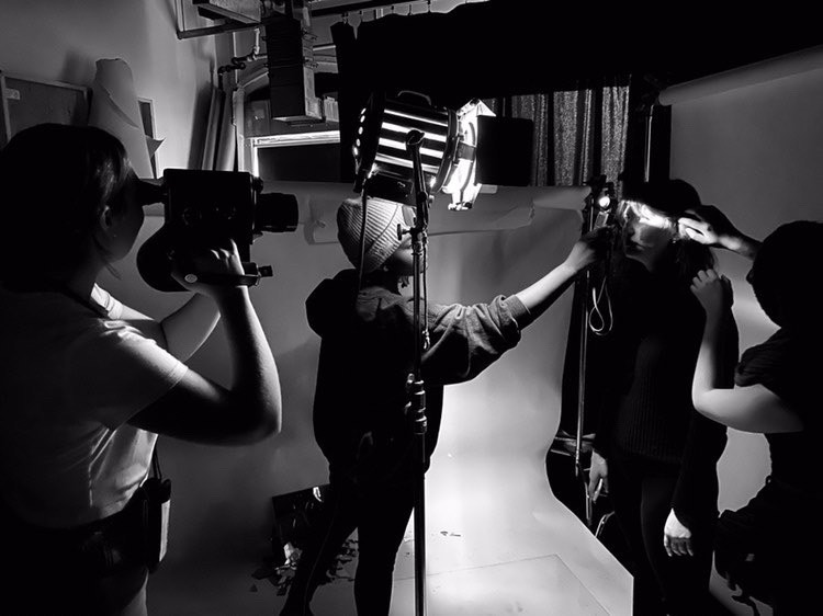 Black and white behind the scenes photo of a film crew setting up a shot. Someone on the left is holding a camera while a person in the middle is helping someone else with the lighting