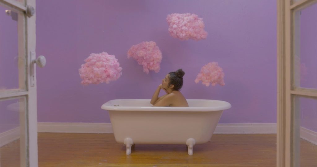 Still image from Domenica Garcia's film. A naked person sits in a bahtub in a purple room with 4 pink clouds on the walls