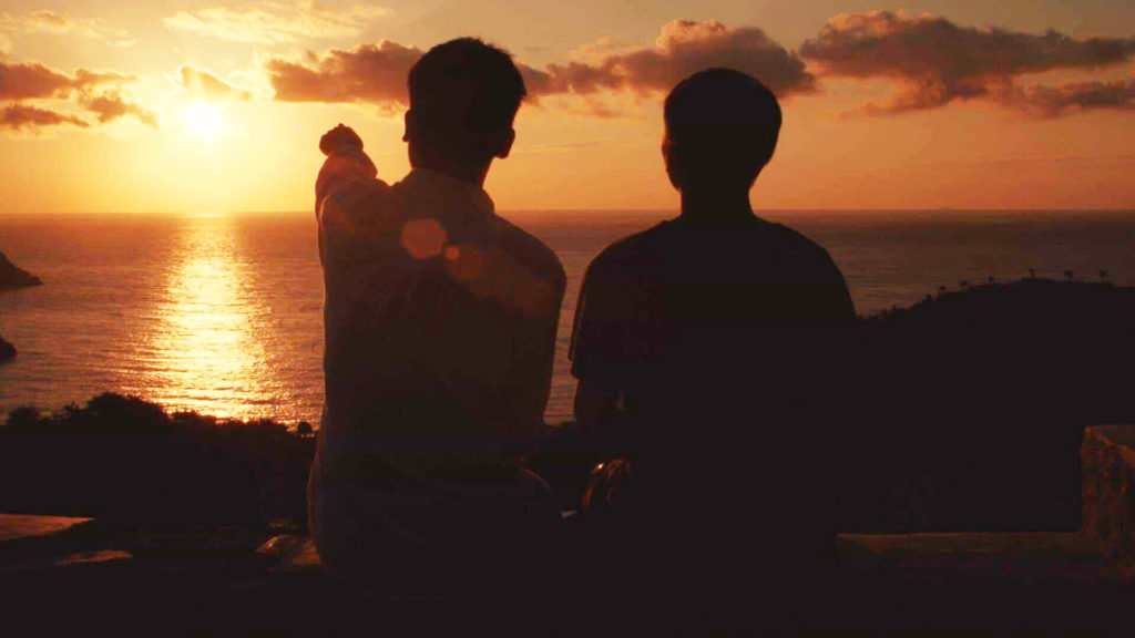 Still image from Meicen Meng's film with two young boys sitting on a hill over a body of water watching the sun set. one of them is pointing at the sun.