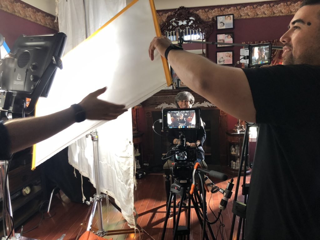 Behind the scenes of an interview. The interview subject sits in front of a camera smiling while two crewmembers set up a chimera lightbox