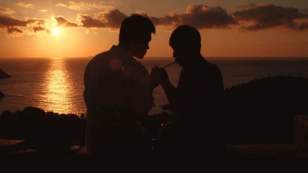 A man lights the cigarette for another man while they sit on the edge of a hill enjoying the sunset over a body of water. 