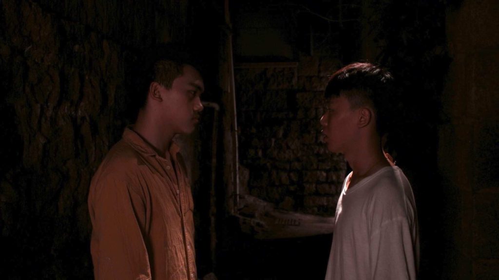 Two men stare intensely at each other, surrounded by blackness. One has a collared button up shirt on and the other a white t shirt.