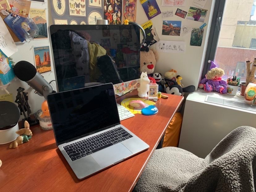 Work from home set up with computer laptop in the SVA Ludlow dorm room. A computer monitor and a laptop sit on a desk surrounded by trinkets and a microphone.