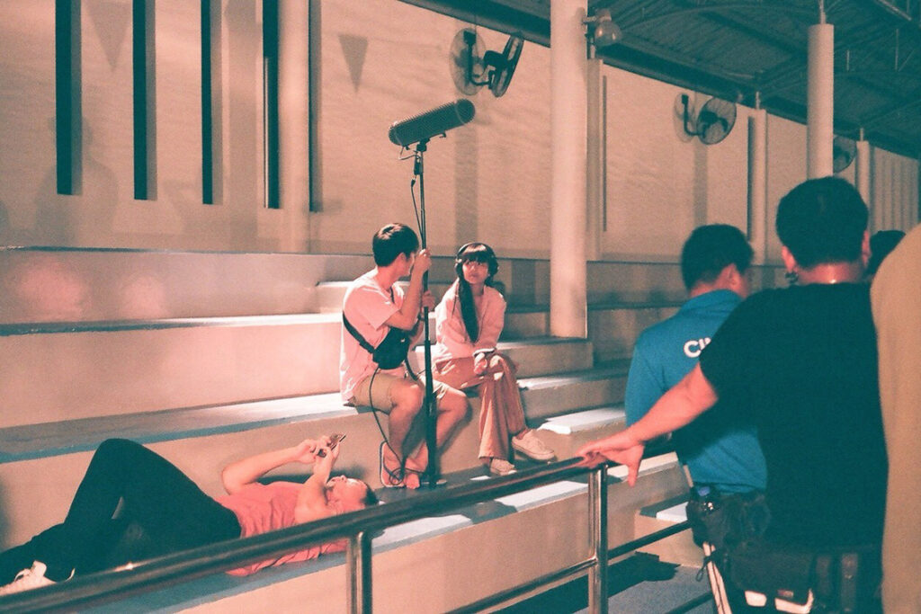 Crew on set of Bouquet Napusorns film. A sound mixer holding a boom pole sits next to another person with headphones while a thirs person lays next to them. Three other people stand off to the side with their faces away from the camera