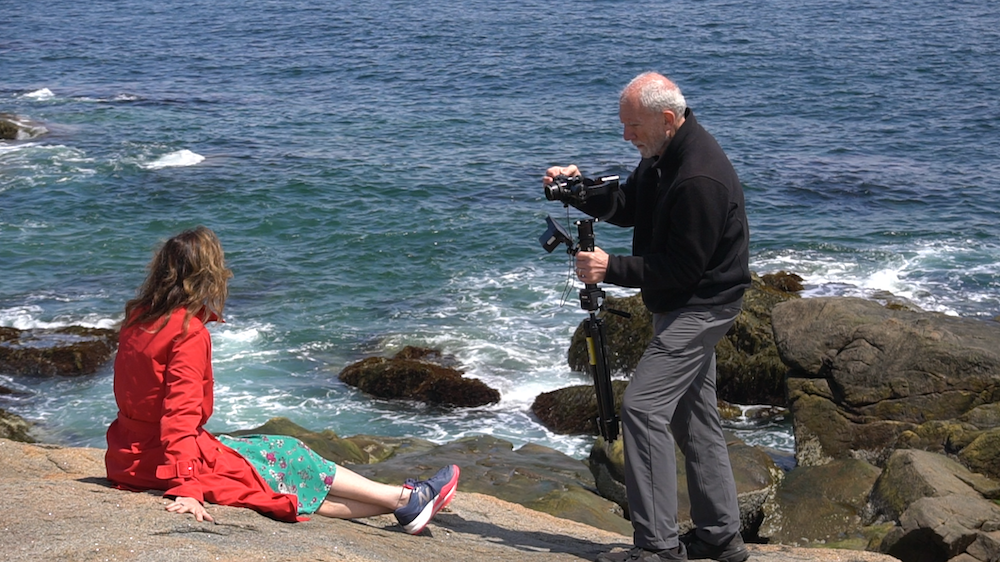 Behind the scenes photo from frank vitale's film which shows frank and a person in a red jacket by the shore of a beach as Frank sets up a camera.