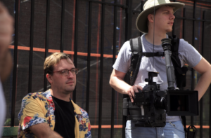 Alumnus Joe Ralko on his film set in a hawaiian shirt, sitting down next to his cameraman holding the camera.