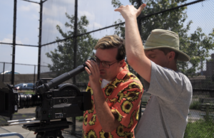 Behinds the scenes on Joe Ralkos film as he looks into a camera viewfinder and a crewmember stands behind him blocking the sun.