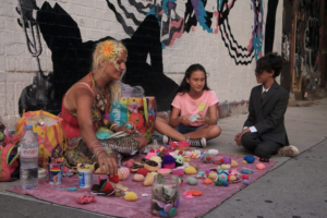 Still image from Joe Ralko's film. Shows a person sitting down outside on a blanket with various trinkets in front of them and two younger people sitting cross legged next to them.