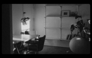Black and white still image from Gonnie Zur's film showing an empty room with minimal decor. An empty desk with a potted plant is on the left and an almost empty bookshelf with another potted plant to the right.