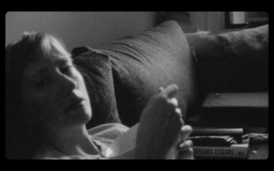 Black and white image from Gonnie Zur's film. Shows a person laying down on a couch, slightly out of focus with harsh shadows on their face.