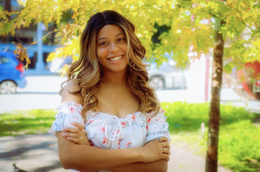 Rianne Pyle looks into the camera with her arms crossed while standing outside on a sunny day.