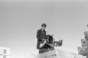 Black and white photo of Person standing on rooftop looking down at a film camera being operated by another person whose face is obscured by the camera