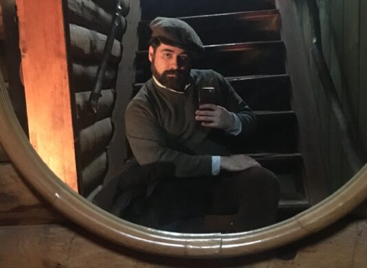 Sean Dermond taking a mirror selfie on set in front of a large oval mirror, wearing a hat and warm clothing.