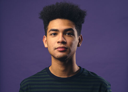 Blog writer Ahmari Ly-Johnson stands in front of a purple background with a sweatshirt on looking directly into the camera.