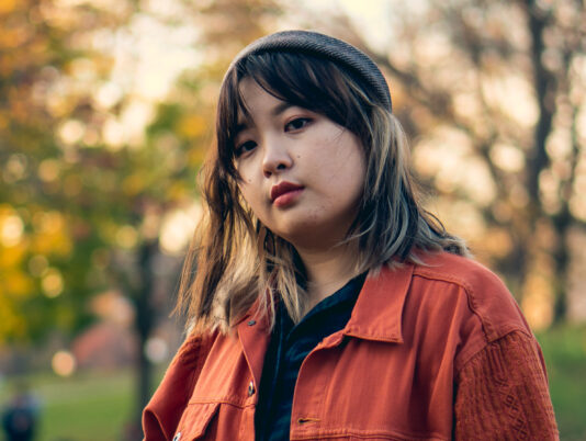 Student Yoko Chen wears a hat and an orange jcaket while standing outside. A blurred background of trees and grass can be seen behind them.