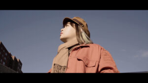 Still from Yoko Chen's short essay film shows Yoko wearing an orange jacket, hat and beige scarf while looking up as a blue sky surrounds them.