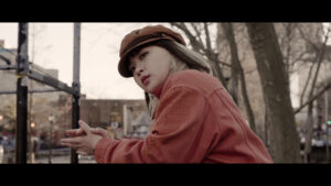 Still from Yoko Chen's short essay film shows Yoko wearing a hat and orange jacket with their left side facing the camera and their face looking at the camera with a thoughtful expression