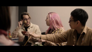A still from Yoko Chen's short essay documentary which shows three people gathered around a table using chopsticks to eat while looking at one another with happy expressions