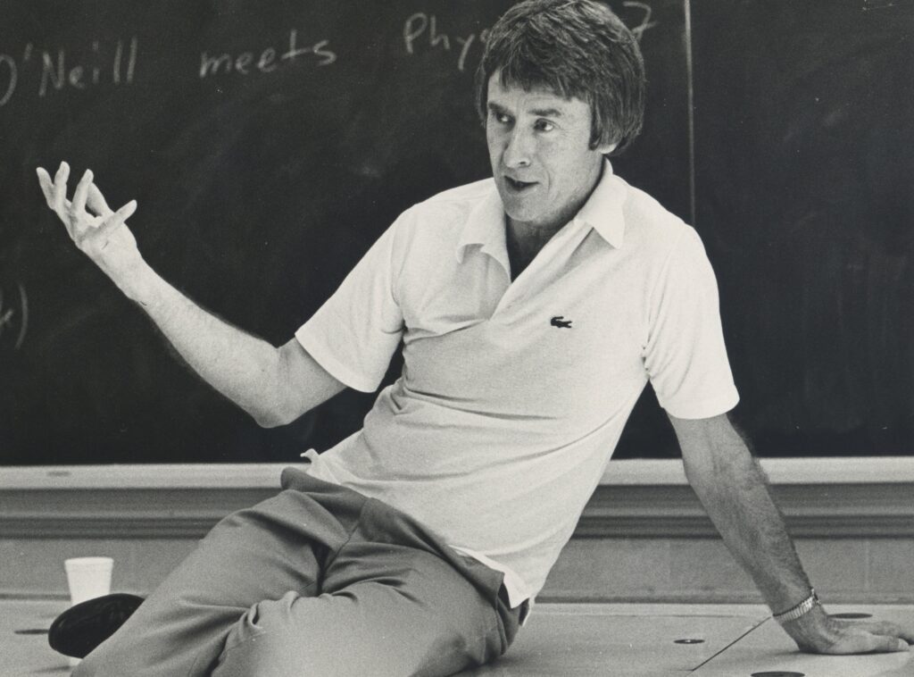 a teacher in a classroom sits comfortable on a desk in front of a blackboard gesturing to people off camera