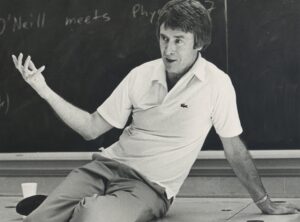 Image of a person in wearing lacoste polo shirt sitting casually on a desk in front of a chalkboard.