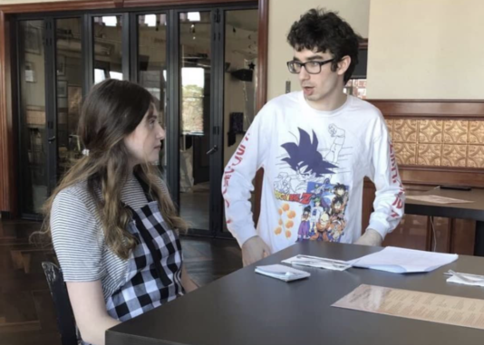 Student Jack Murtha stands next to a person sitting at a desk, speaking with them on the set of their short film.