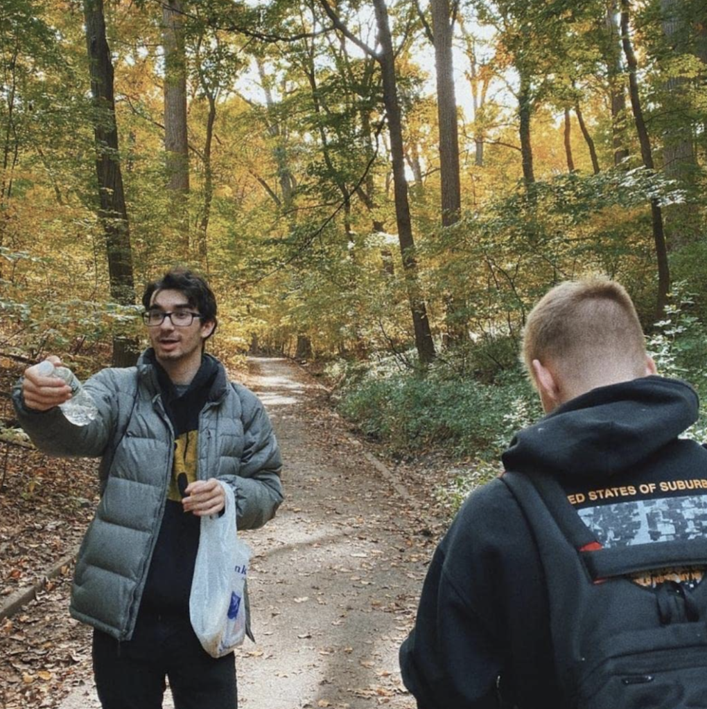 two people standing outside in the woods on a trail. One of them has their back turned to the camera, the other facing the camera holds a plastic bag and a water bottle while gesturing to someone off camera.