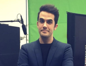 Will Henry looking directly into the camera wearing a sharp looking blazer with a background of film equipment (green screen, Boom microphone) behind him