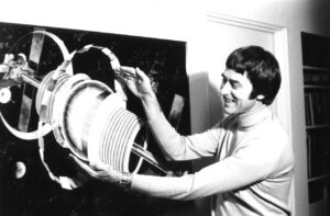 Black and white image of a person in a turtleneck holding their hands in front of a picture of a spaceship in outer space.
