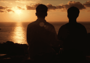 Still image from Meicen Meng's thesis film shows two men sitting on a cliff overlooking water and a sunset
