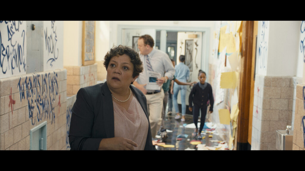 Still image from Dan's Film Showing a woman in a school hallway looking confused while children and other faculty walk behind them.