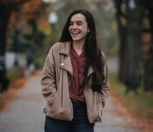 Student Annie Fleisch stands on a street surrounded by fall trees and orange leaves, wearing a jacket and smiling.